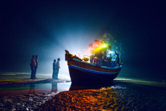 Boats bedecked with lights returning from a pilgrimage wait in the shoals for the tide to return so that they can head back home to their village. 2019 © Arko Datto